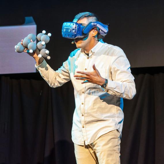 omeone on stage wearing a VR headset, holding an atom diagram.