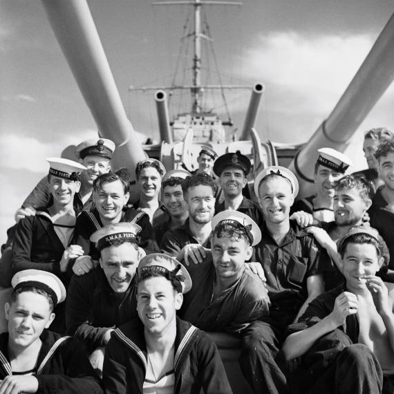 group of men in navy uniform on HMAS Perth smiling for a photo sitting under the ships guns