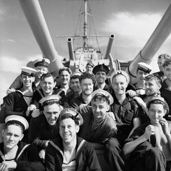 group of men in navy uniform on HMAS Perth smiling for a photo sitting under the ships guns