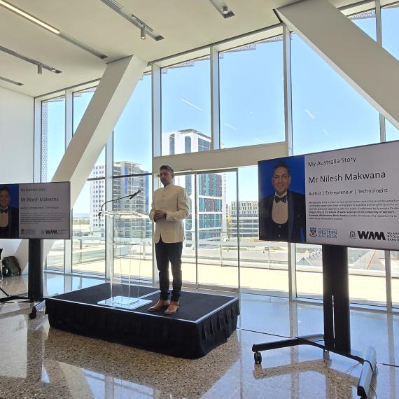 A person stands on a podium flanked by 2 screens, large windows behind them