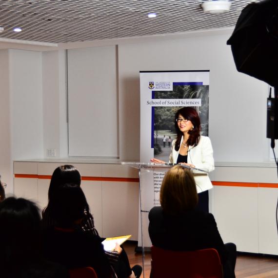 A person in a white jacket stands at a lectern in a white room and presents to a seated audience in front of them.