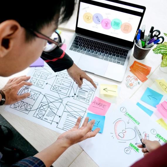 Image shows a person working at a desk with a laptop. They have paper in front of them which has post it notes helping them to work through a design process.