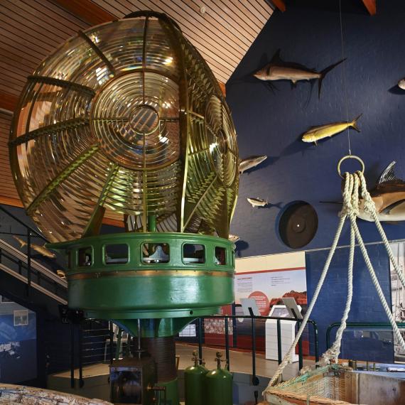 A large Fresnel lens in a green frame, part of a museum exhibit with marine-themed decorations and informational panels in the background.