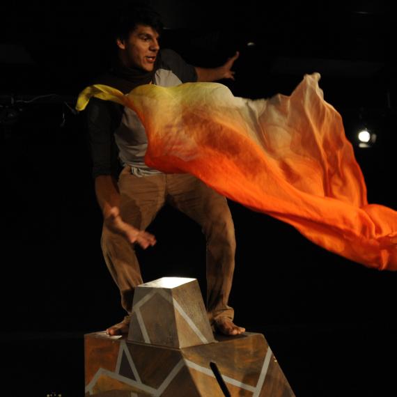 Yirra Yaakin Theatre Company presents Kaarla Kaatijin, a performance for young people about Noongar country and the importance of fire knowledge.  This scene features a performer standing  high on staging and reacting to a flame-coloured waft of fabric in a dynamic action moment.