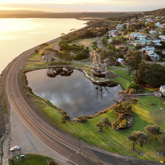 An aerial shot of a verdant coastal landscape, featuring a wooden ship beached by a small lake, and a road curving alone the coastline.