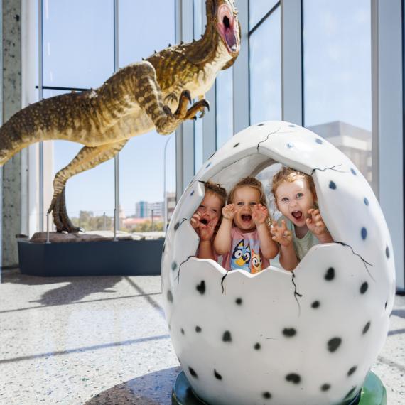 Three children inside a cracked dinosaur egg with a dinosaur model behind them in a museum.