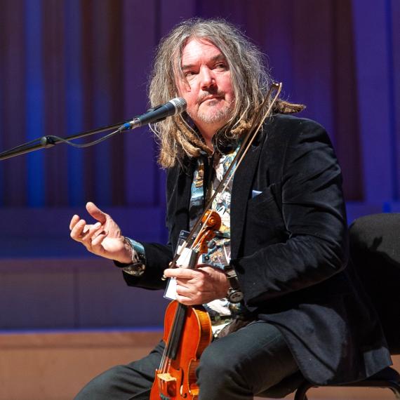 a person sitting on a stoll with a violin in hand talking
