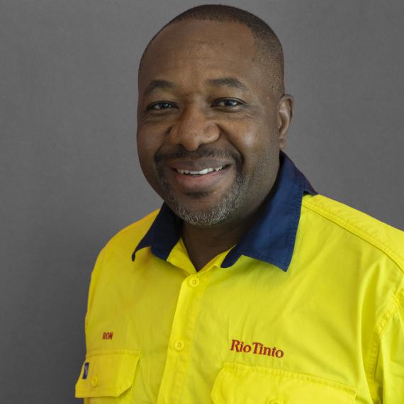 A man wearing a bright yellow shirt with "Rio Tinto" logo smiles against a gray background.