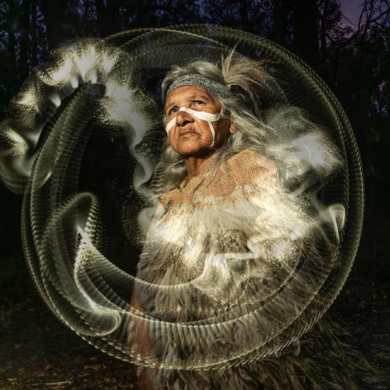 an Aboriginal Elder with traditional ochre on her face and arms looks up. She wears clothing made of emu feathers. There is a white circular halo and dust around her