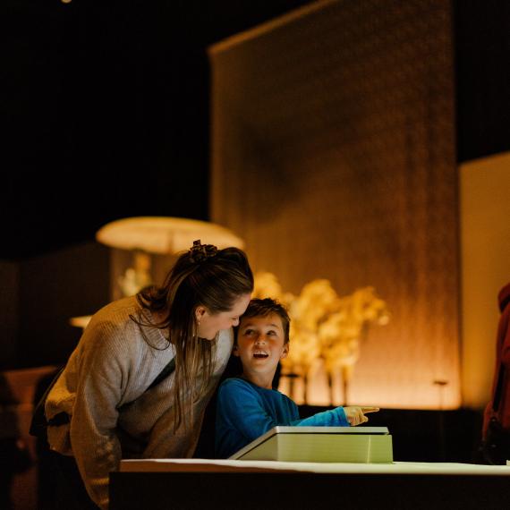 An adult and a child looking at an illuminated display table in a dimly lit room with warm lighting