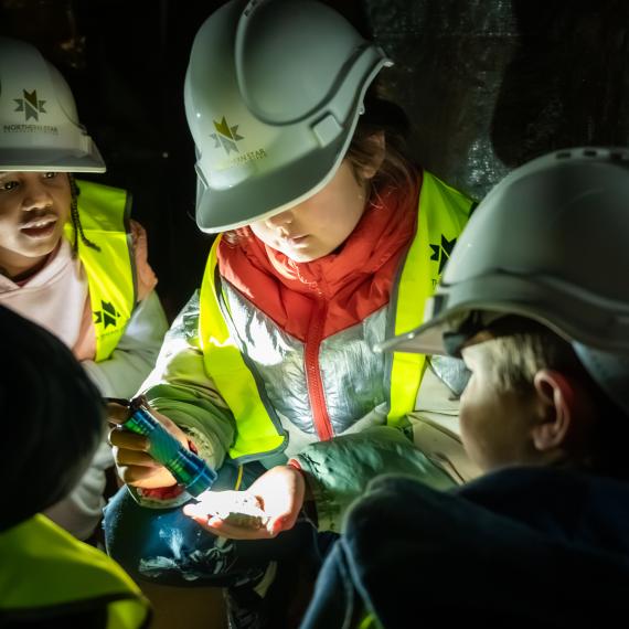 Children wearing hard hats and safety vests, examining an object in dim light.
