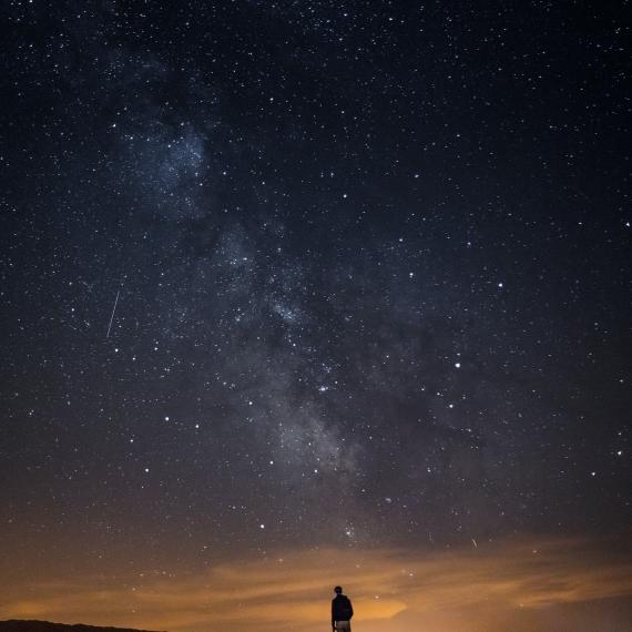 Image shows a dark, starry night sky. A person is silhouetted against an orange horizon. 