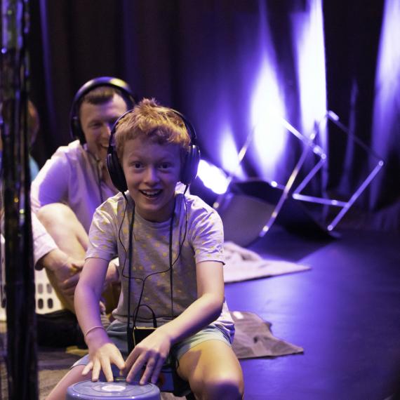 Smiling child wearing headphones squatting on blanket with upturned bucket in front of them. Smiling adult behind them also wearing headphones.