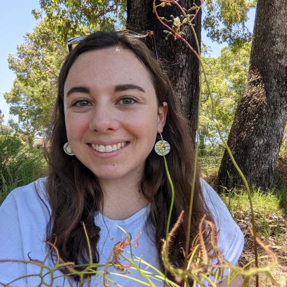 Image shows a close up of Laura Skates the presneter for this Meet the Museum session. She has long dark hair ans wearing a grey T-shirt. Bushland is in the background 