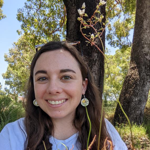 Image shows a close up of Laura Skates the presneter for this Meet the Museum session. She has long dark hair ans wearing a grey T-shirt. Bushland is in the background 