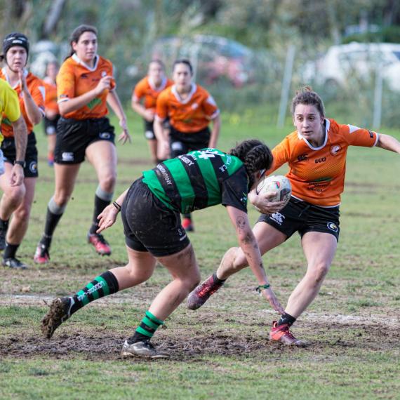 Image shows two competing teams of women playing rugby. ONe team has an orange uniform and the other a green and black striped uniform. The playing field is muddy. 