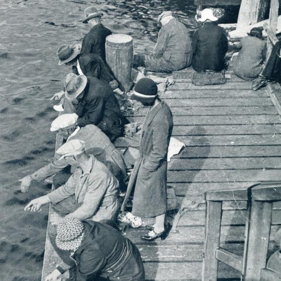 An archival photograph showing a group of people are leaning over a dock and pointing in the water 