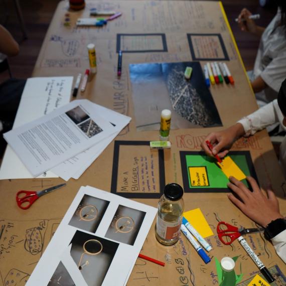 The image shows a long table covered in brown paper and various materials that students are using for a design activity.