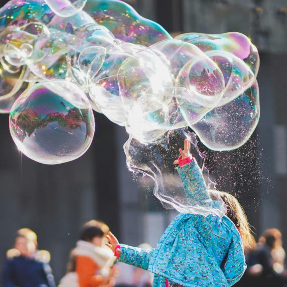 Child in blue jacket reaches into the air to touch giant bubbles floating past