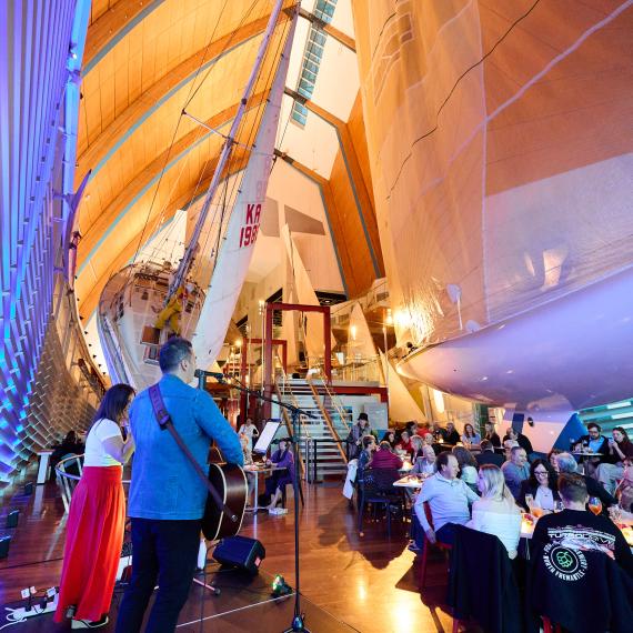 people seated on tables, performers on a small stage in the foreground with yachts in the background