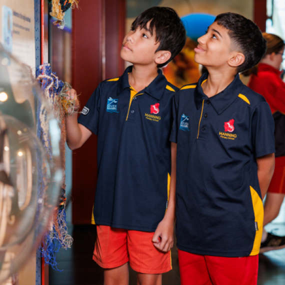 Two students in navy and yellow school uniforms are closely examining a textured display featuring woven materials and fibers. The exhibit appears to showcase traditional or natural elements, possibly related to cultural heritage or environmental themes. The setting is indoors, with bright lighting and other students visible in the background, suggesting an educational excursion or museum visit focused on interactive learning.