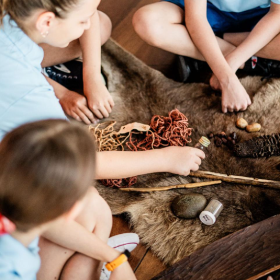 A group of students sits in a circle around a display of cultural artifacts arranged on an animal hide. The collection includes natural fibers, wooden sticks, stones, and small containers, representing traditional tools and materials. The setting suggests an interactive learning activity focused on Indigenous knowledge, history, or cultural practices, encouraging hands-on engagement and discussion.
