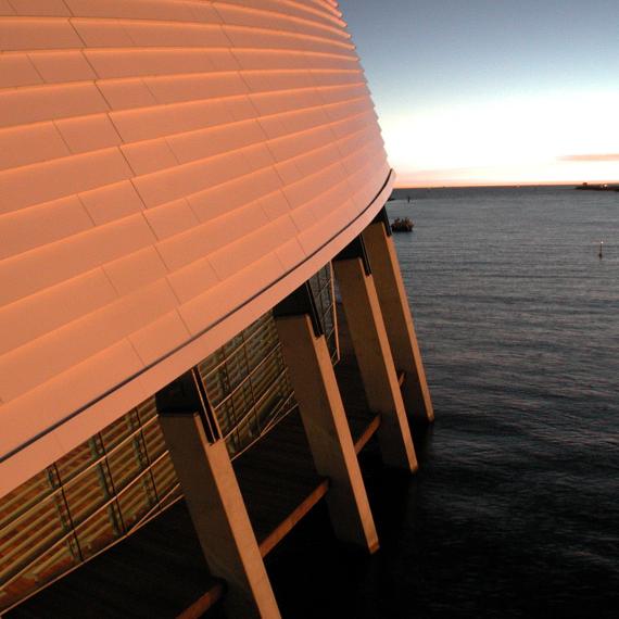 Half the photo pictures the outside of the museum - its curved roof, windows, boardwalk and supporting concrete beams. The other half pictures a river. There a few clouds in the distance that look orange in colour as the sunsets.