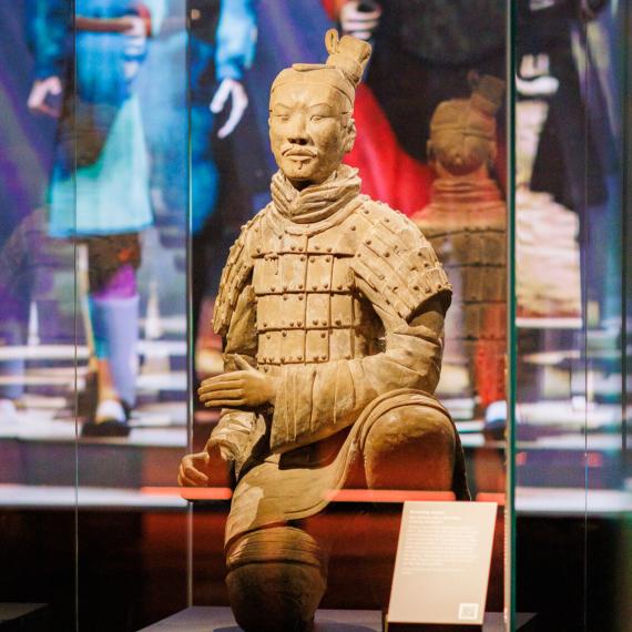 Terracotta warrior statue displayed in a glass case with an information plaque, colourful figures visible in the background.