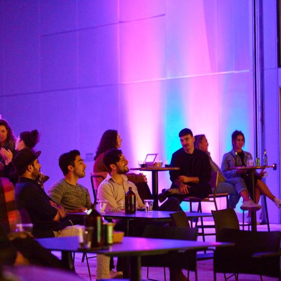 People seated at tables in a dimly lit room with purple and blue lighting, engaged in conversation at a social event.