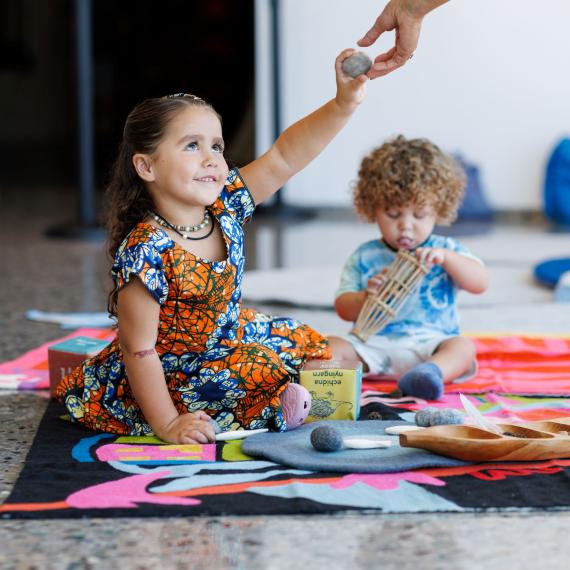 Two young children sitting on a mat - someone passing something to the young girl.