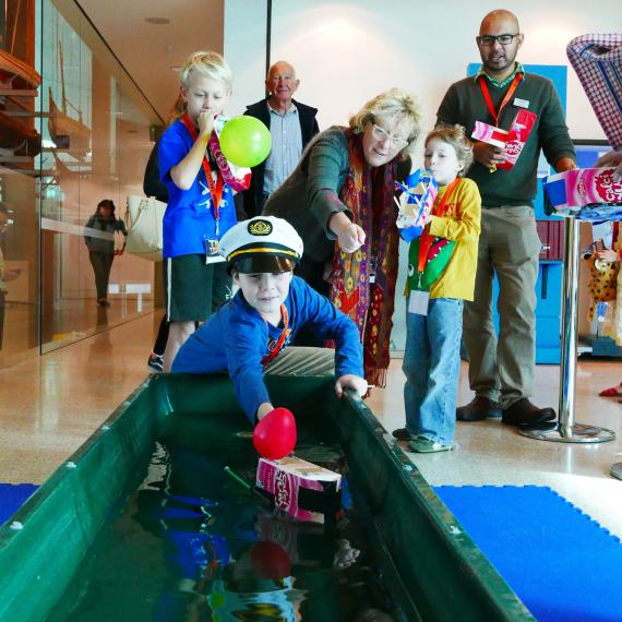 a young child wears a sailors hat and pushes their milk carton boat down a long indoor paddle pool. other children and adults watch on with happy smiles 