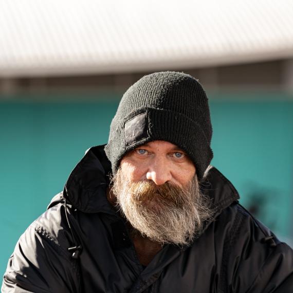 an adult with a beard wearing a beanie stares straight ahead into the camera