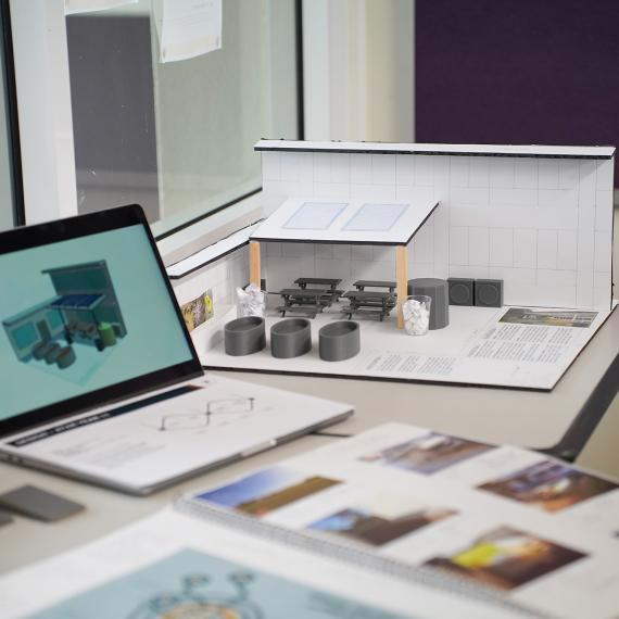 Architectural model of a small building displayed on a desk beside a laptop showing a 3D design, with sketches and photos spread around.