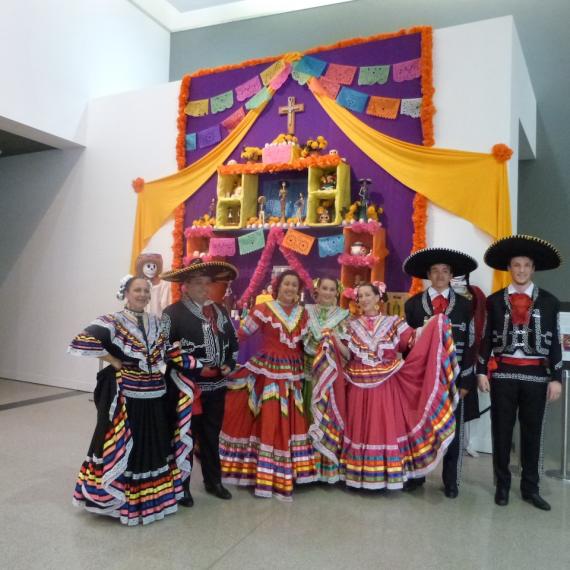 A group of people in colourful traditional costumes standing in front of a decorated Día de los Muertos altar inside a gallery space.