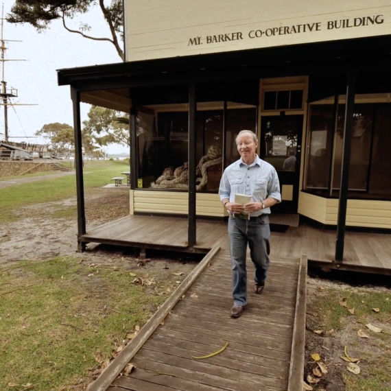 Person walking from the Mt Barker Co‑operative Building, with a historic sailing ship and waterfront in the background.