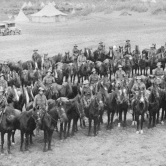 Black-and-white photo of mounted troops lined up on horseback, with tents of a military camp in the background.