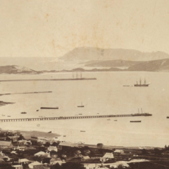 Sepia-toned panoramic view of a coastal town and harbour, with long jetties, anchored ships, and distant hills across the water.