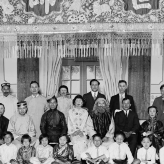 Black-and-white group portrait of adults and children posed in rows inside an ornate interior with decorative curtains and wall panels.