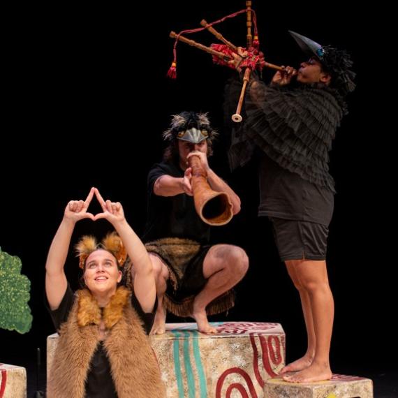 Yirra Yaakin Theatre Company present Djinda Kaatijin. This production image shows a trio of young Aboriginal theatre performers, one seated with her hands in a star formation above her head; one crouched nearby on a stage block playing didgeridoo, and the third performer standing on a stage block playing the bagpipes.
