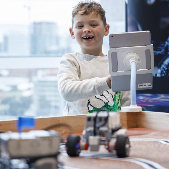 Young child standing in front of an iPad smiling. Robots on table in foreground