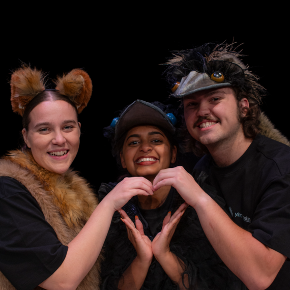 Three performers from Yirra Yaakin' 2026 production of Djinda Kaatijin, shown together in costume (Emu, Crow and Dingo) all smiling and making a loveheart shape with their six hands together.