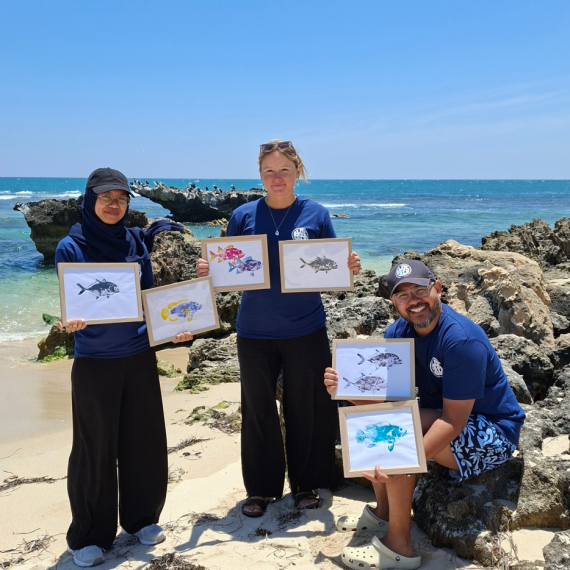 three people are on a beach with the ocean behind them. One person is sitting on a rock, the others are standing' All art holding some artwork showing fish. 