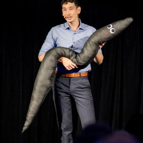Man on stage holding large worm-like soft toy