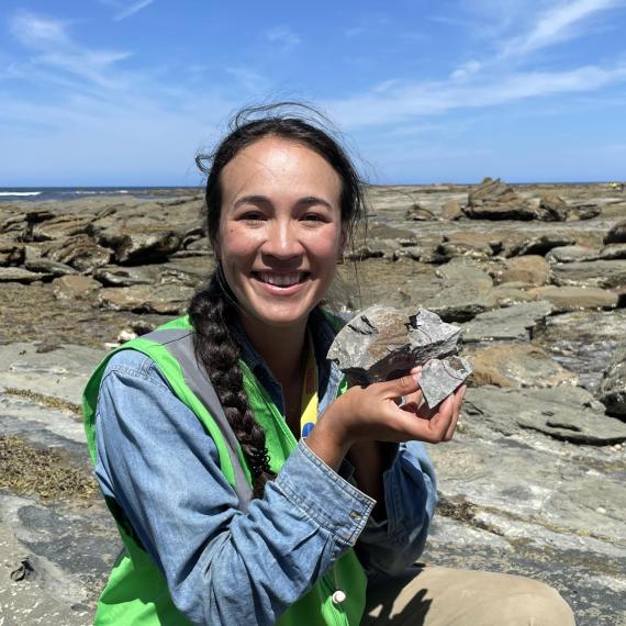 an adult person outside in a rocky landscape holds up a rock with some fossils inside the rock