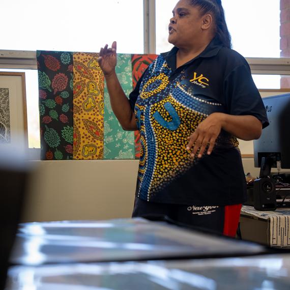 a person stands and is talking facing the side and in the background artworks are being displayed