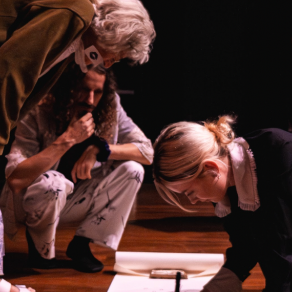 A woman with blonde hair and a black jumper in leaning over a white piece of paper on the floor holding a pencil. a Woman with short grey hair and an olive green jumper in leaning down towards the paper while another person with brown hair in crouching in the background