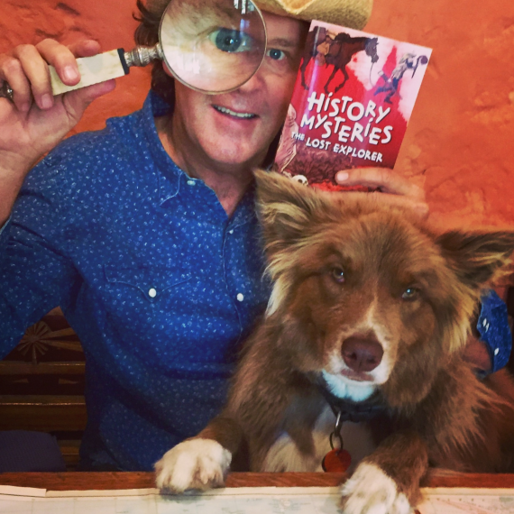 A man with brown hair, a blue shirt and a straw cowboy hat is holding a large magnifying glass in front of one eye. he is holding a book with a red cover titled History Hunters: the Lost Explorer and a map spread out on the table in front of him. On his lap is a brown and white dog  