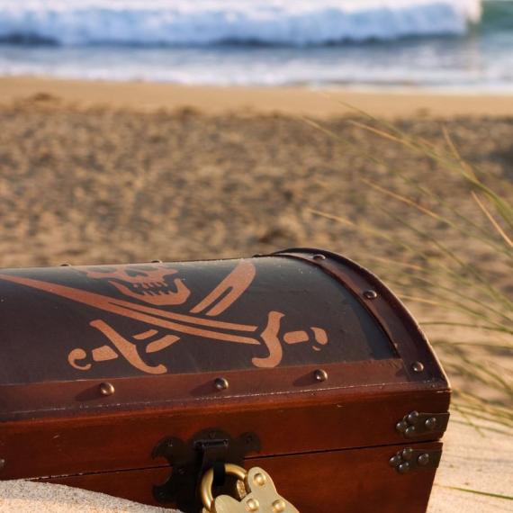A treasure chest buried in sand on the beach with crossed swords and a skull showing on the lid, linked to the Treasures of the Deep event for Totally Lit 2026 festival.