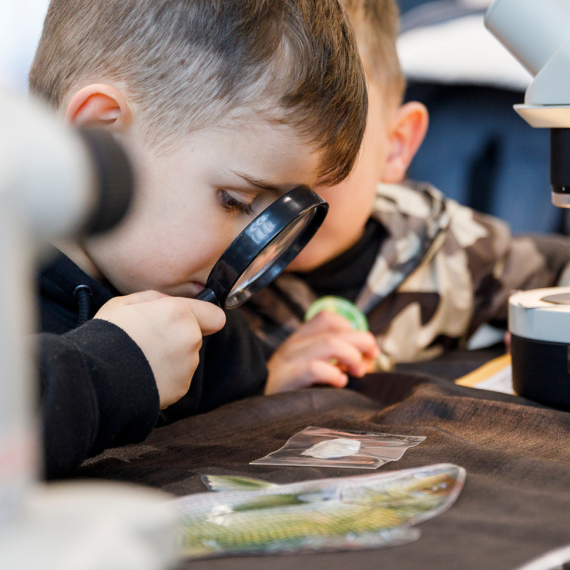 Kid looking through magnifying glass at object with telescope behind him