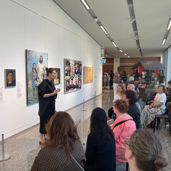 A woman with a black shirt and brown hair pulled into a bun is standing in front of a wall covered in paintings. She is speaking to a group of people sitting on stools in front of her. 